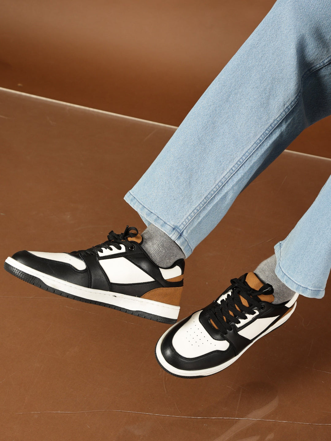 Black and white sneakers worn with light blue jeans on a brown floor.
