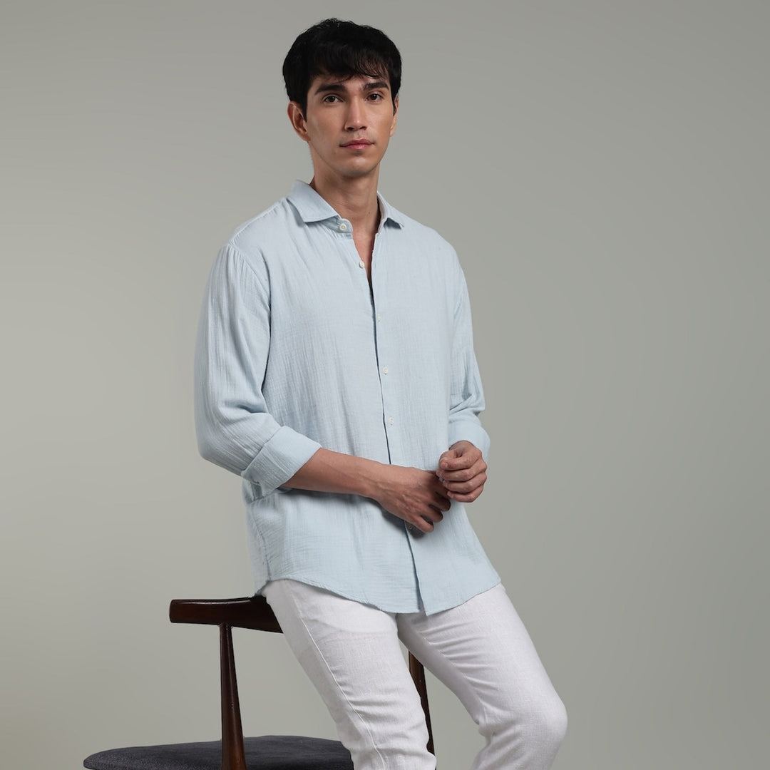 Man wearing a light blue shirt and white pants leaning  on a chair against a gray background