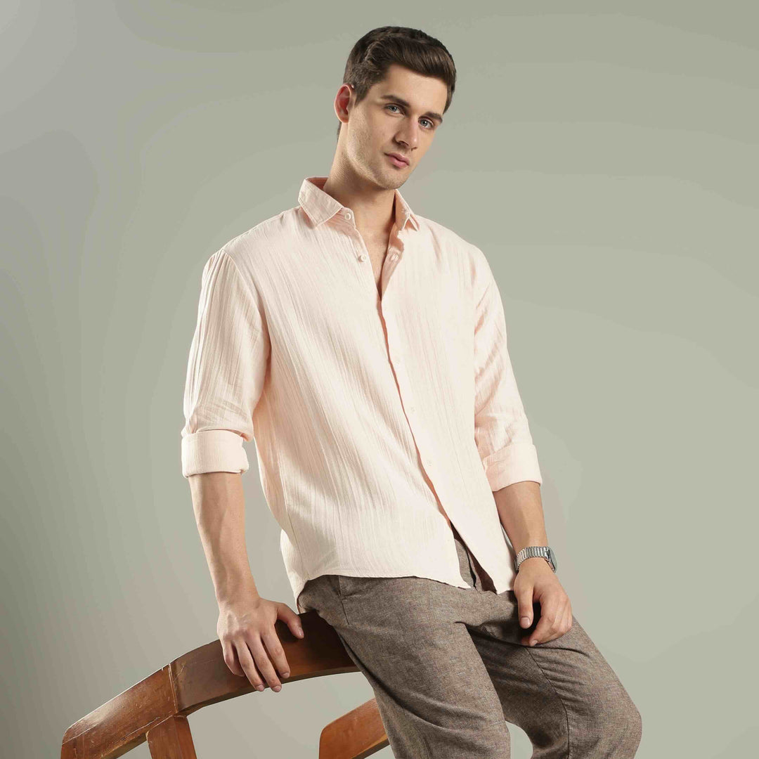 Man wearing a light pink shirt and gray pants sitting on a wooden chair against a plain background
