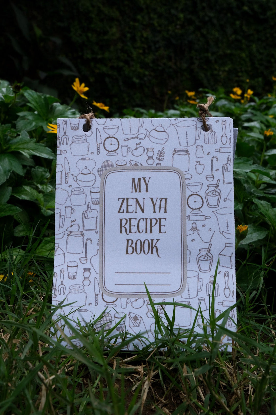 My Zen Ya Recipe Book with illustrated cover placed on grass and yellow flowers – wellness drink recipe book from Zen Ya Rakhi gift box.