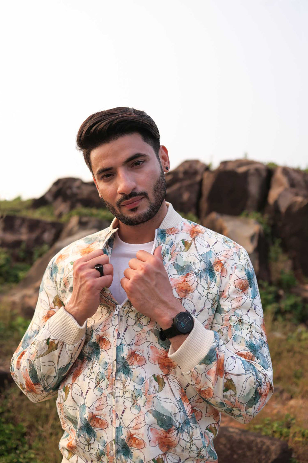 A man standing against a white background wearing a multicolored floral hemp bomber jacket with a round neckline and a front zipper.
